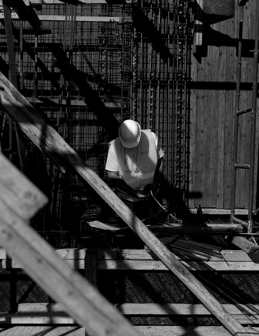 Un operaio con casco e gilet ad alta visibilità legge un progetto in cantiere, tra ferri e tavole di legno